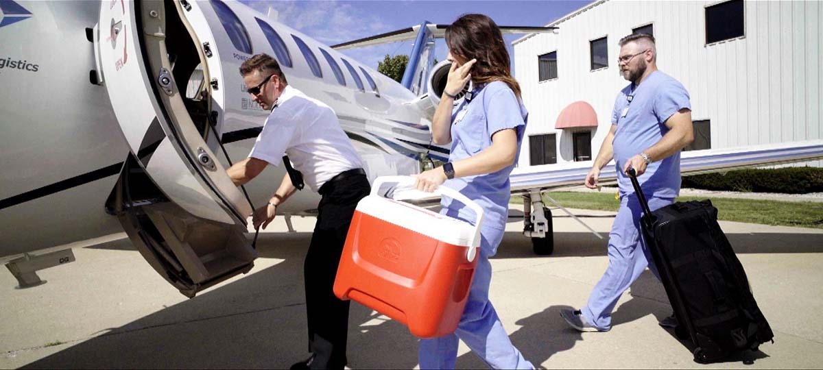 Doctors in blue scrubs carry a red cooler and a black suitcase aboard a plane.