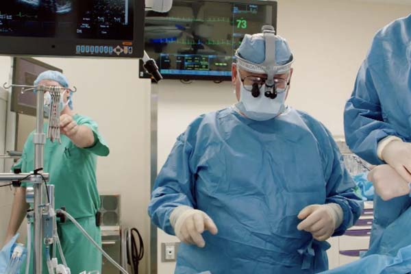 Dr. Patrick McCarthy dressed in scrubs by an operating table. Play Video.