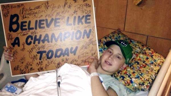 A young boy lays in a hospital bed, wearing a Notre Dame baseball cap, holding a signed Believe like a champion today sign.