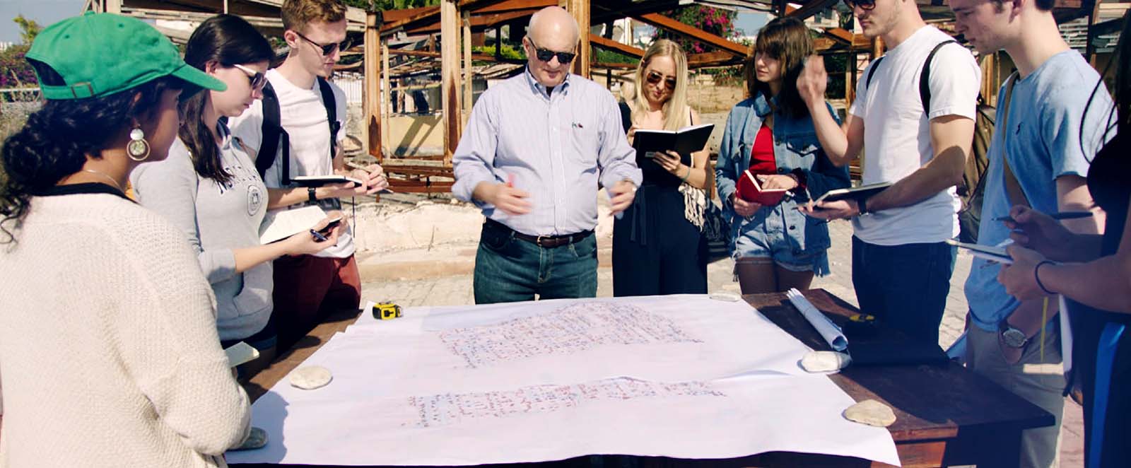 A professor and group of students stand around a table with architectural sketches on top.