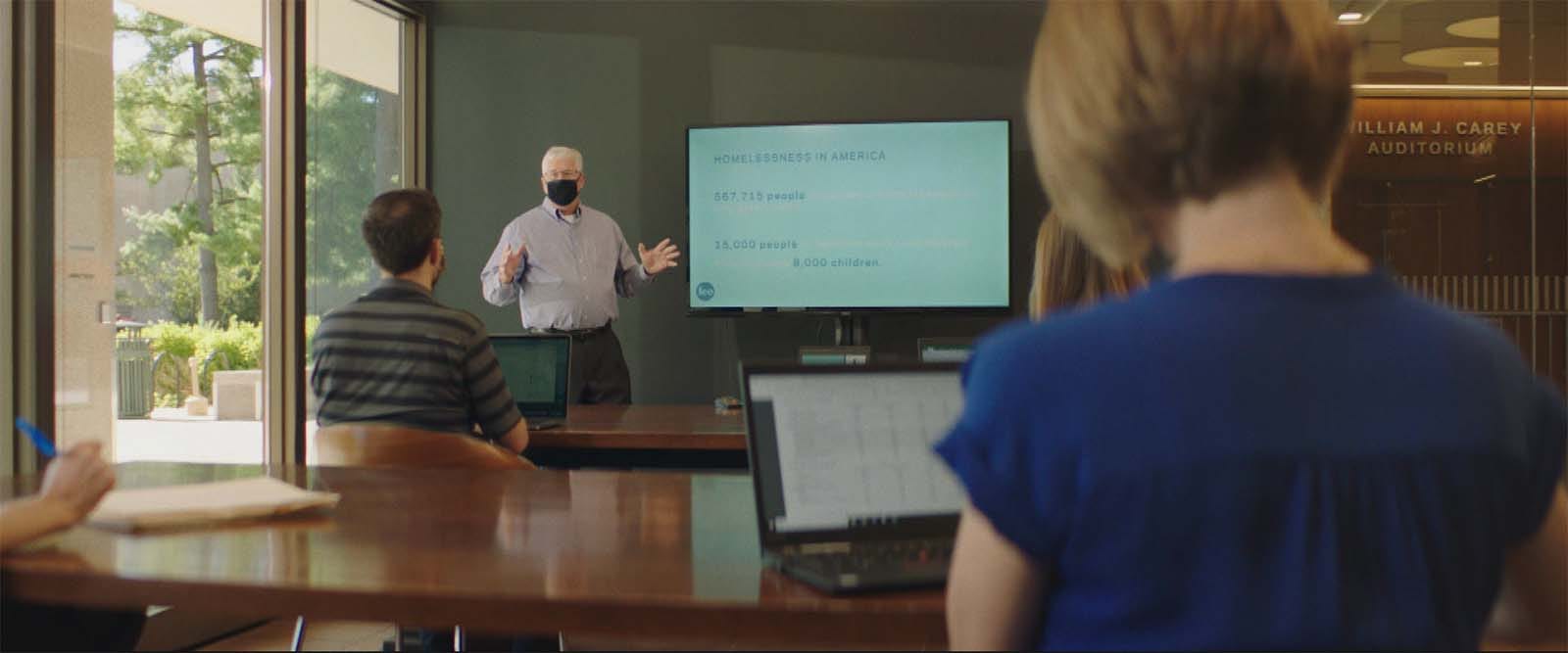 A man in a face mask presents in front of screen to a classroom of students.