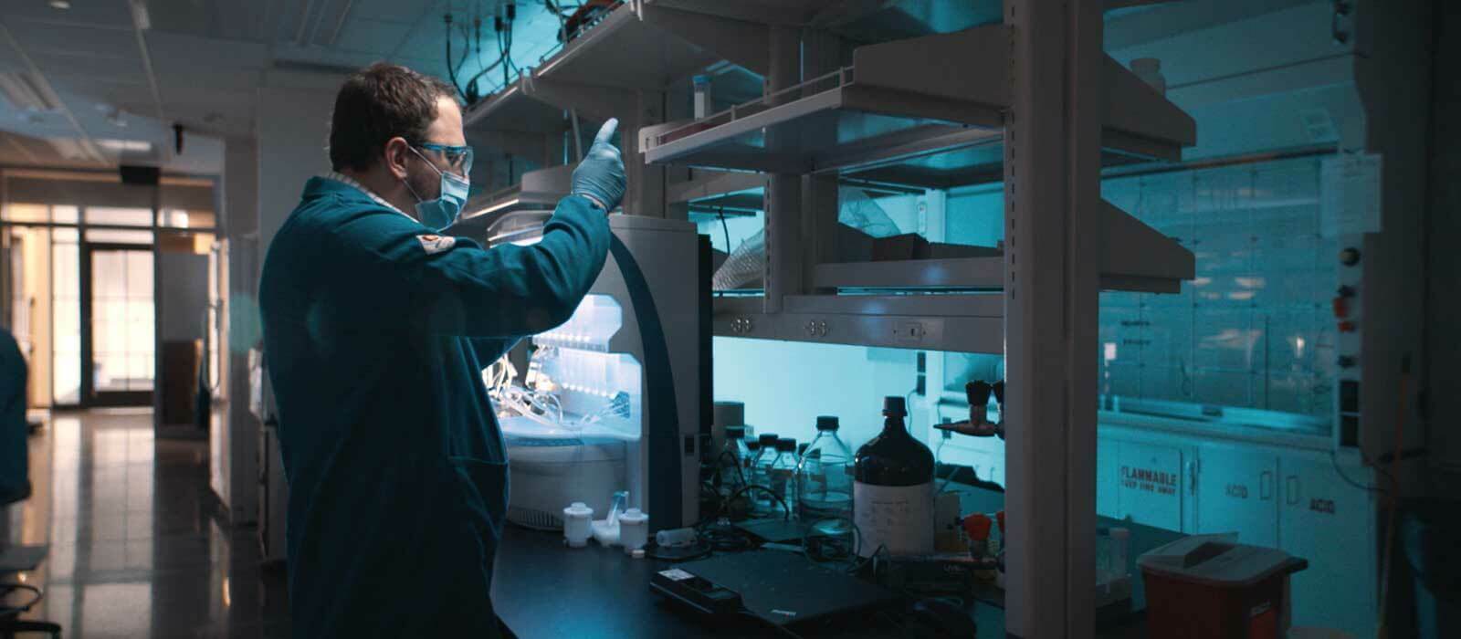 A researcher stands in a dimly-lit lab, filling a test tube using a syringe.