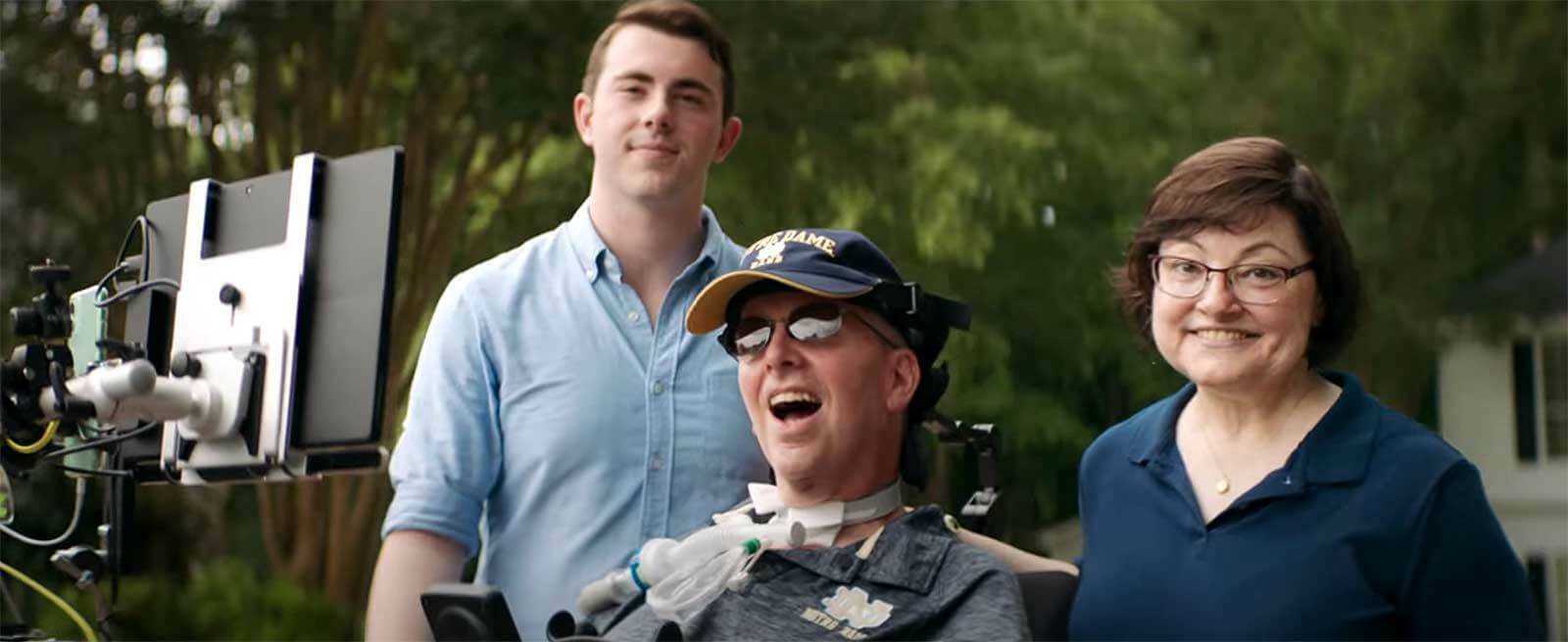 Two men and a woman are outside, blurred trees in the background. One man is in a wheelchair and the other two stand next to him.