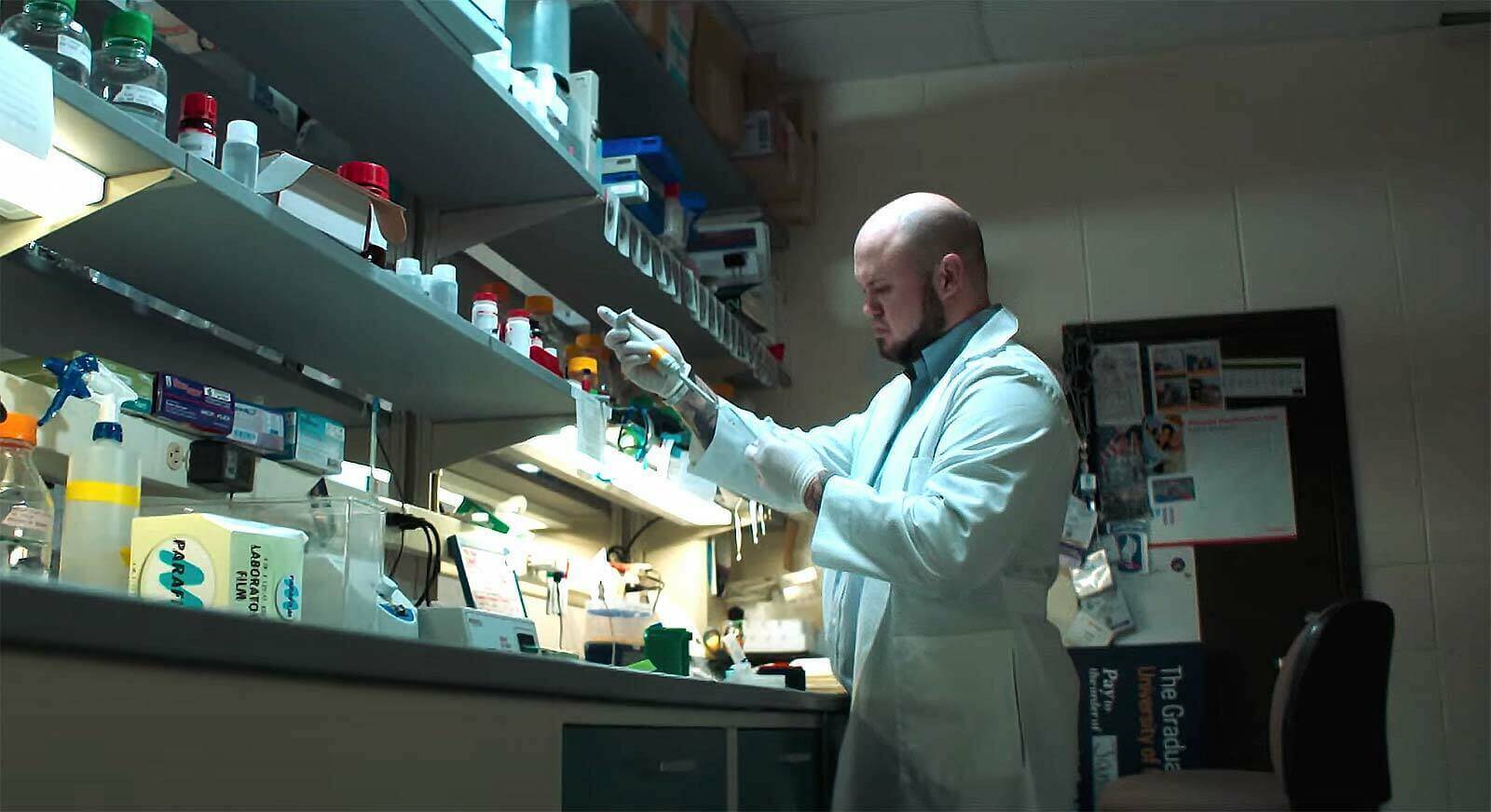 A man standing inside of a lab, wearing a white lab coat and gloves holds a syringe and test tube.