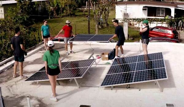 A group of six students work on top of a roof, moving solar panels in place.