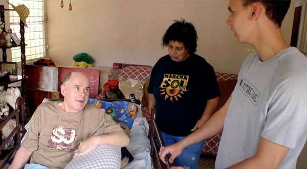 A male student speaks with two residents in their home. One man, sitting on a couch, looks up to the student.