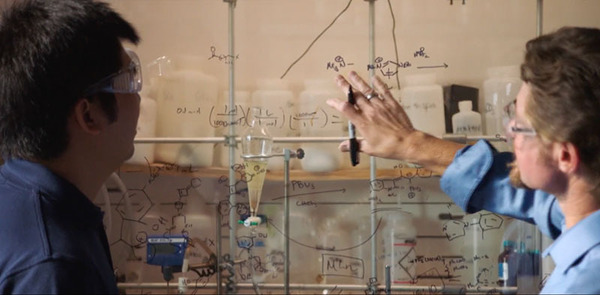 Two male professors work on chemical equations using marker on a window. There are chemicals in glass jars in the background