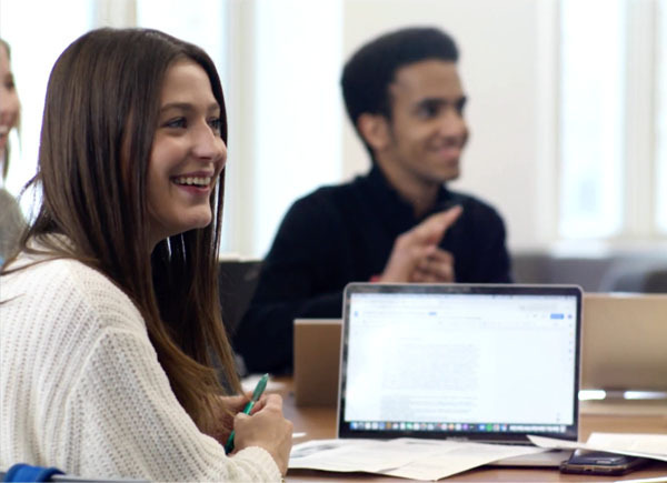 Female student sits in front of laptop with a male student in the background