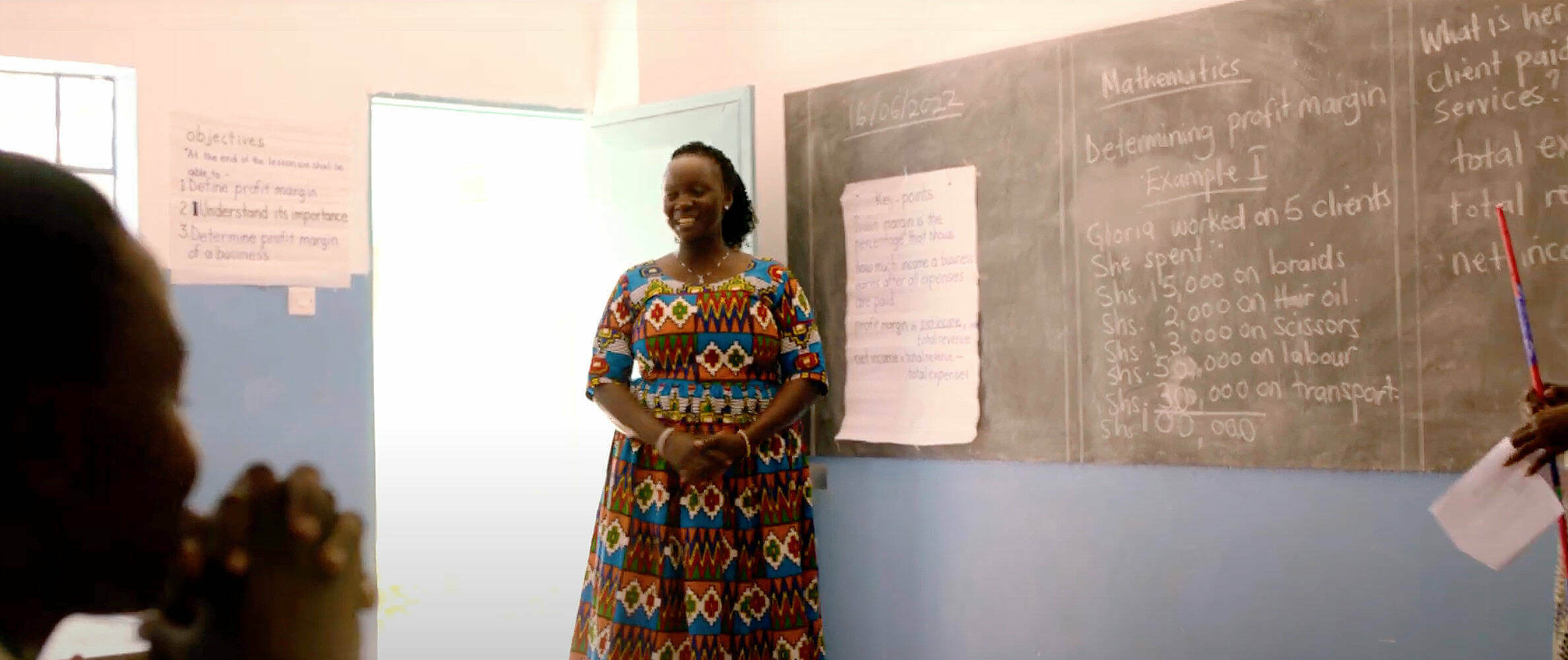 A black woman stands in front of a chalkboard in a classroom.