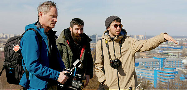 One male holding a video camera instructs two other males.