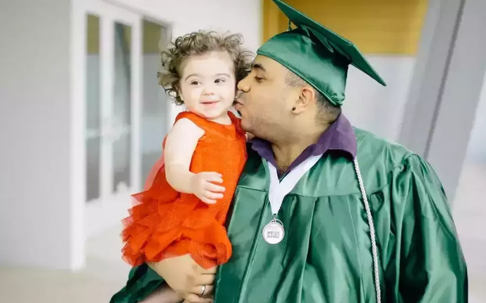 Man wearing cap and gown kisses cheek of toddler in his arms.