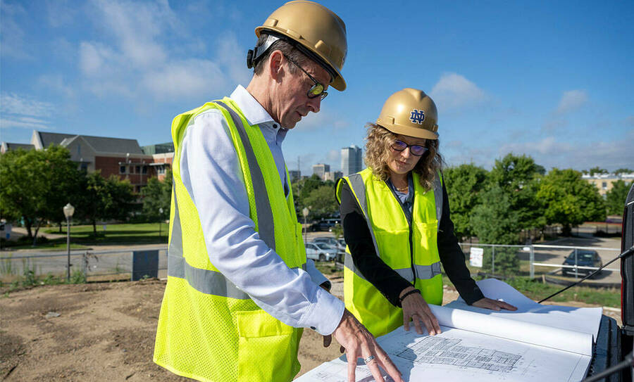 Sarah Mustillo looks over blueprints with a contractor. She's wearing a neon vest and gold helmet