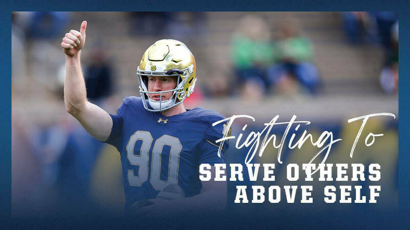 A Notre Dame football player, wearing a gold helmet and a blue jersey with the number 90, gives a thumbs-up on the field. Text overlay reads, &quot;Fighting To Serve Others Above Self.&quot;