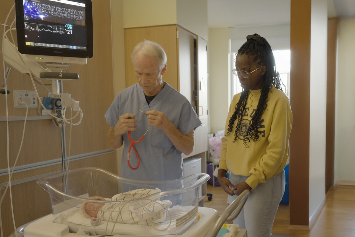 A doctor in blue scrubs holds a red stethoscope, preparing to examine a newborn lying in a hospital bassinet. A young woman wearing a yellow Notre Dame sweatshirt stands beside them.