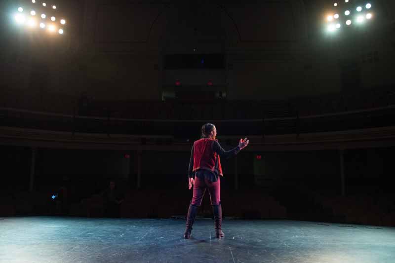 A person stands on a stage, lit by spotlights, with their arm extended while performing. 