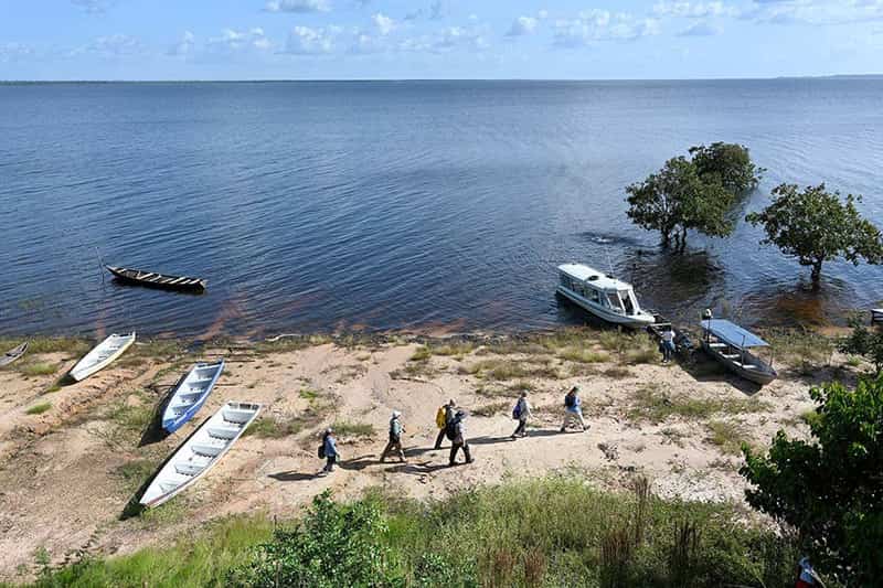 People walk along a sandy shoreline near canoes and motorboats with a large body of water in the background.