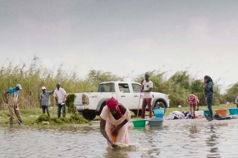 A group of people are standing in muddy water, with one woman in the foreground washing clothes in a plastic basin. 