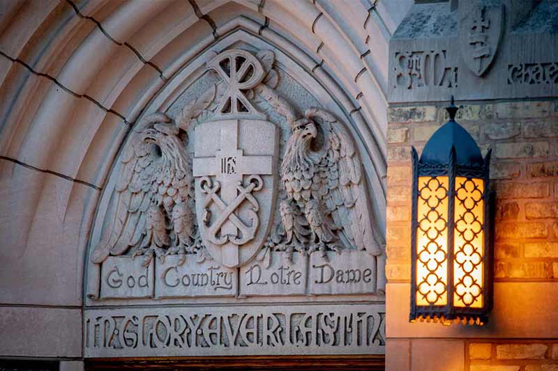 A Notre Dame coat of arms carved into stone with the motto ‘God, Country, Notre Dame’ is illuminated by a decorative lantern.
