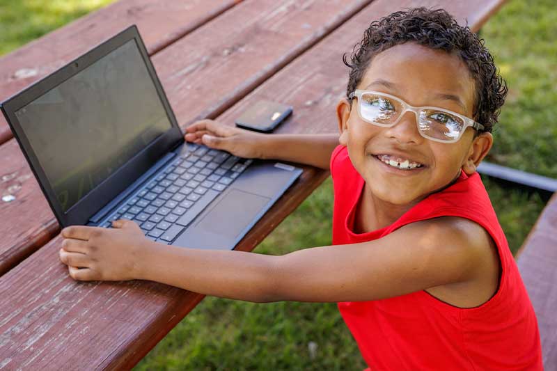 A smiling young boy with curly hair and glasses sits at a picnic table, with his hands on an open laptop. 