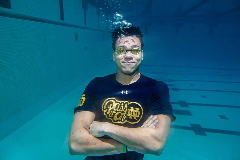 A man underwater in a swimming pool wears goggles and a dark shirt with the Notre Dame logo, smiling with his arms crossed. 