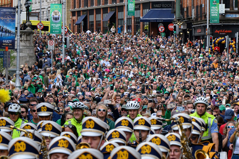 A large crowd of people gathered on a city street in Dublin, Ireland. The foreground features members of the Notre Dame marching band, identifiable by their uniforms, including hats with the Notre Dame logo. The background is filled with a dense crowd, with some individuals holding up phones to take pictures or videos. 
