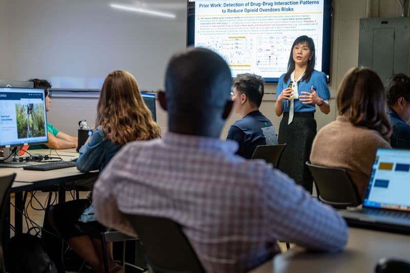 A woman in a blue shirt and black skirt gestures as she presents to a classroom of students. 