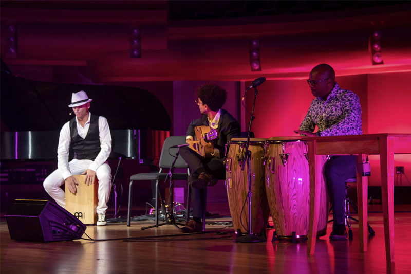 Three musicians perform on a stage bathed in pink light.  One, wearing a white fedora and black vest, plays a cajón.  Another plays an acoustic guitar. The third sits at a table playing congas. A grand piano and other equipment are visible on stage.