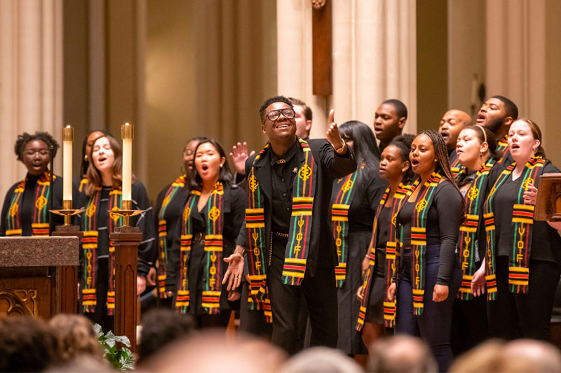 A choir performing in the Basilica of the Sacred Heart. The choir members are wearing black shirts and stoles with a black, red, and green pattern. 