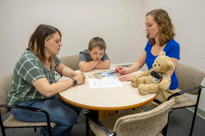 Two women and a boy sit around a table looking at papers and stickers. A teddy bear with a device sits on the table next to the woman in blue.