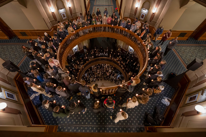 Students hold lit candles on the balconies and grand staircase of the Main Building at the University of Notre Dame.  The view is from above, looking down the spiral staircase.