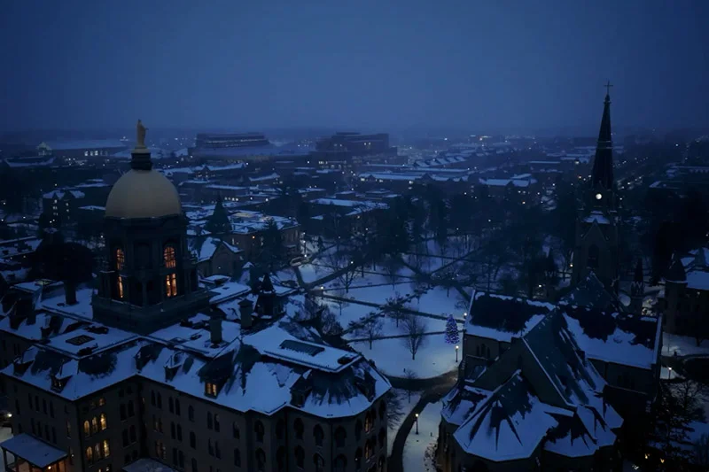 Snowy winter night at the University of Notre Dame. The Golden Dome of the Main Building and the spire of the Basilica of the Sacred Heart glow, surrounded by warm lights and a blue-lit Christmas tree.