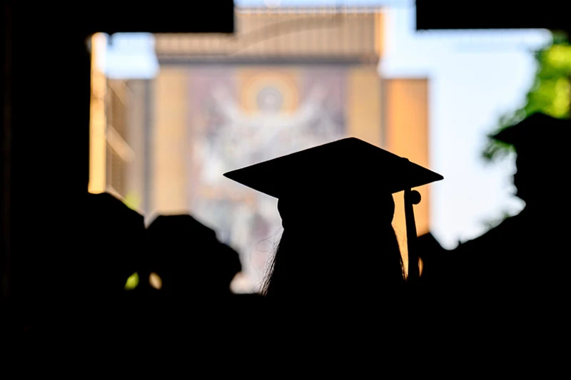 Silhouetted graduates in caps and gowns face the Hesburgh Library's “Word of Life” mural at Notre Dame, seen through an archway.