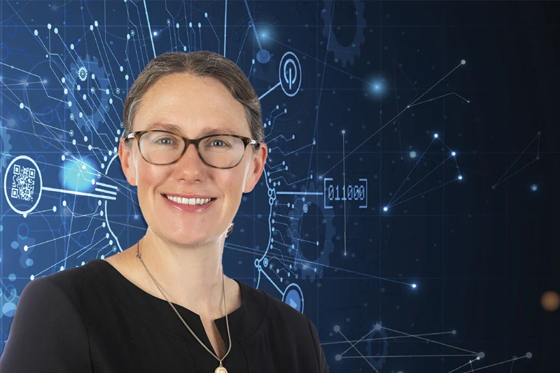 A smiling woman with glasses and brown hair pulled back stands in front of a dark blue background with abstract white digital lines and binary code.