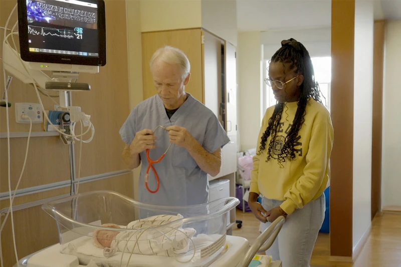 A doctor in blue scrubs holds a red stethoscope, preparing to examine a newborn lying in a hospital bassinet. A young woman wearing a yellow Notre Dame sweatshirt stands beside them.