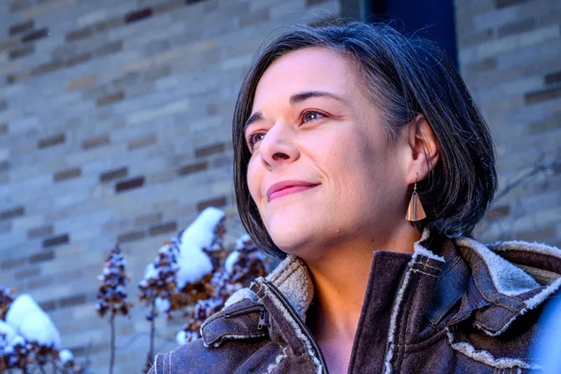 A woman with short dark hair smiles slightly while looking off-camera. She wears a brown, fleece-lined winter jacket. In the background, snow rests on dried plants in front of a tan brick wall.
