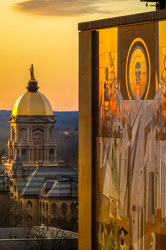 The Golden Dome of the Main Building and the “Word of Life” mural on the Hesburgh Library at Notre Dame are bathed in warm, golden light at sunset.
