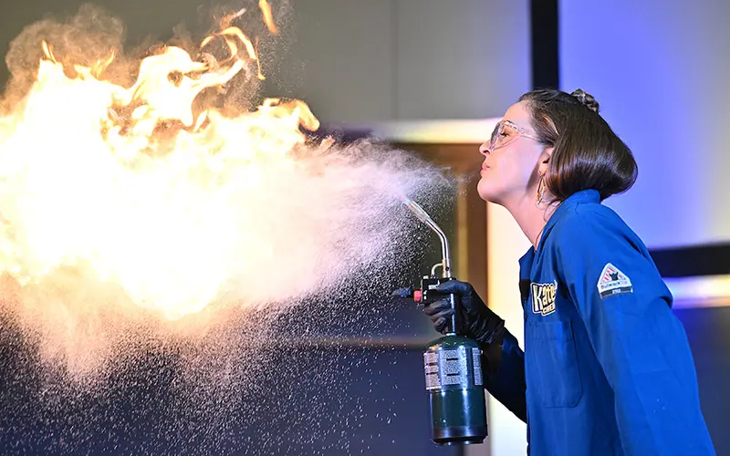 A woman in a blue lab coat, safety glasses, and black gloves aims a small torch at a burst of flames, demonstrating a chemical reaction.	Fine particulate matter hangs in the air around the flame.
