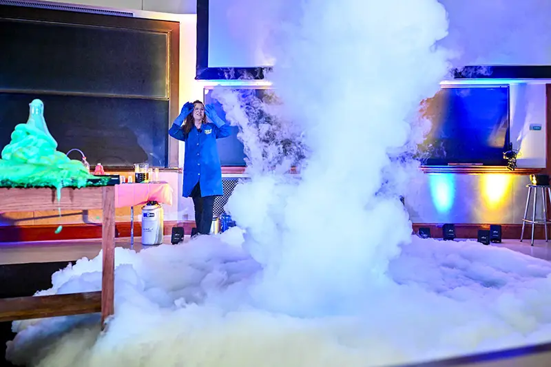 A white female scientist in a blue lab coat reacts to a large white cloud of foam expanding across a classroom floor.	Bright green slime spills over a table to the left. A chalkboard and projection screen are visible in the background. Colorful lights illuminate the scene.
