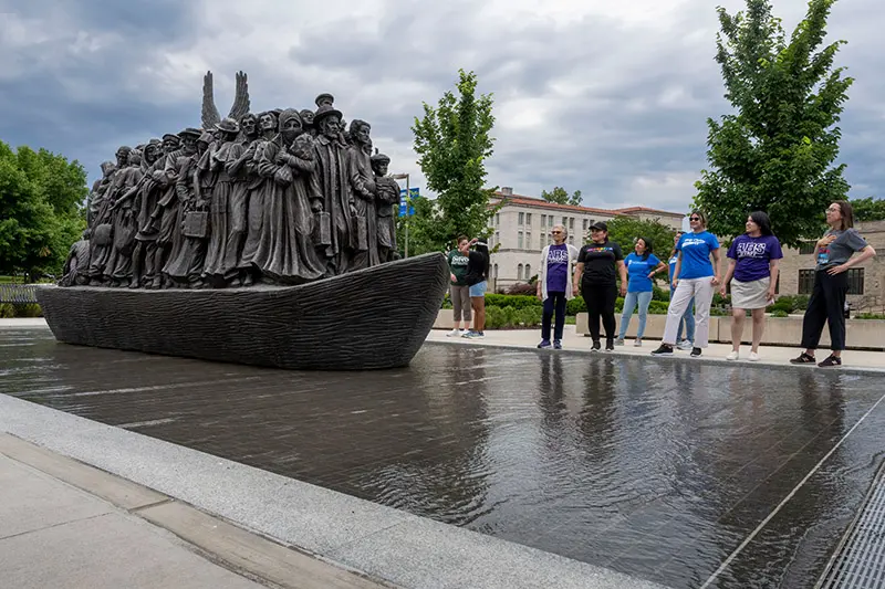 A group of people stand near a large bronze sculpture of immigrants on a boat, symbolizing the journey to a new life.	A partially cloudy sky is above.