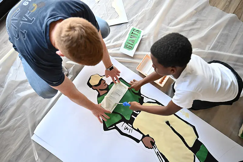 A black boy and a white man kneel over a large piece of paper on a drop cloth, carefully painting a life-sized illustration of a baseball player wearing a green shirt and khaki pants.	The boy uses a small paint roller to apply tan paint.	A small green and white sign reading GIANTS lies nearby.