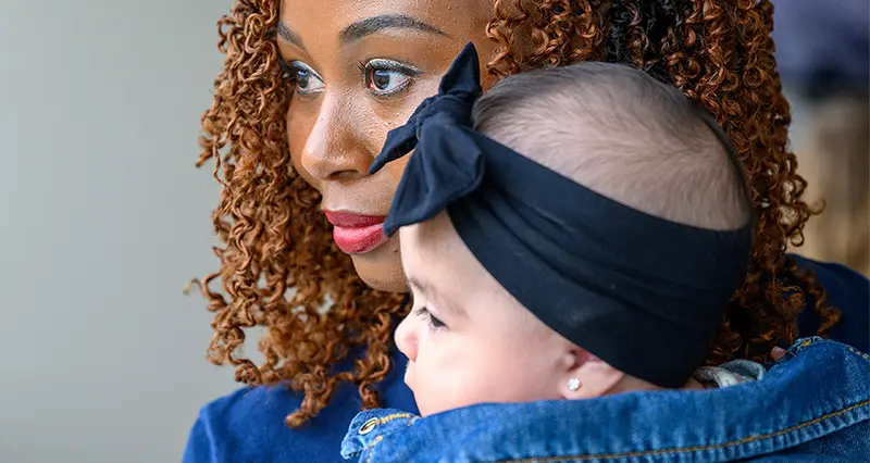 A woman with brown curly hair and red lipstick holds a baby wearing a denim jacket and black bow headband.