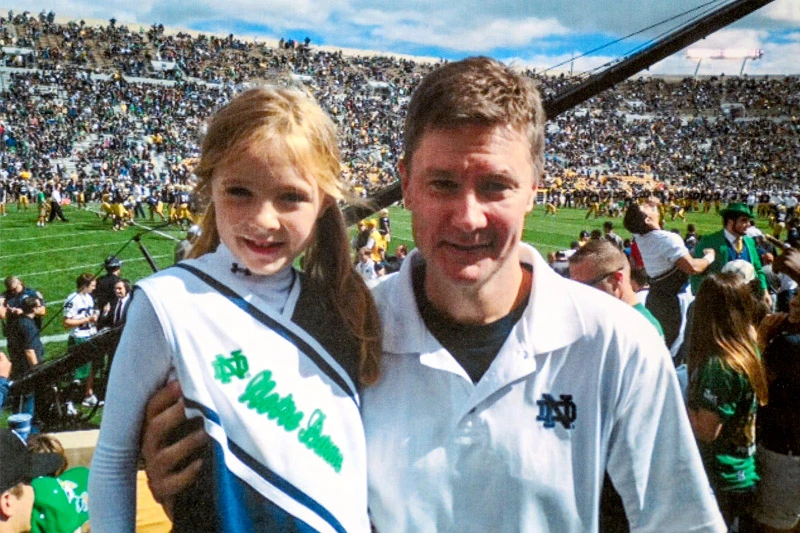 A smiling girl in a Notre Dame cheerleading outfit poses with a man in a white Notre Dame polo shirt in Notre Dame Stadium during a game. The crowded stands and football field are visible in the background.