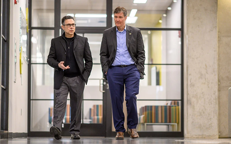 Two men in business attire walk and talk in a hallway with glass-walled rooms. The man on the left gestures with his hand as he speaks.