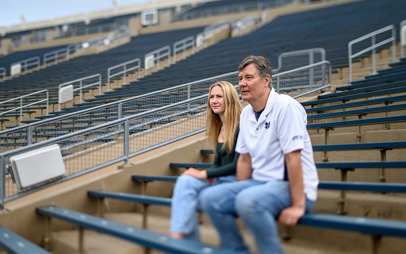 A man and a young woman sit in the empty bleachers of Notre Dame Stadium. The man wears a white Notre Dame polo shirt and jeans. The woman wears a dark green shirt and light-wash jeans. They are looking towards the field.