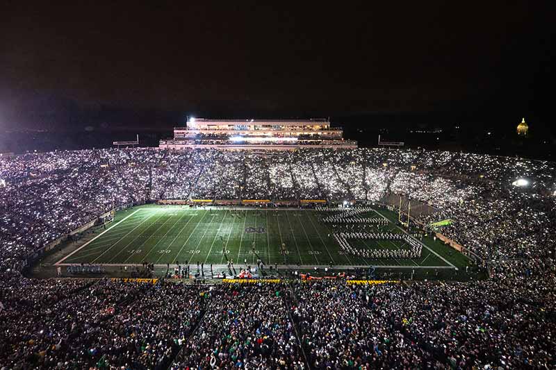 Notre Dame Stadium lit up at night by the fans phone flashlights. In the background, the Golden Dome	glows against the night sky.