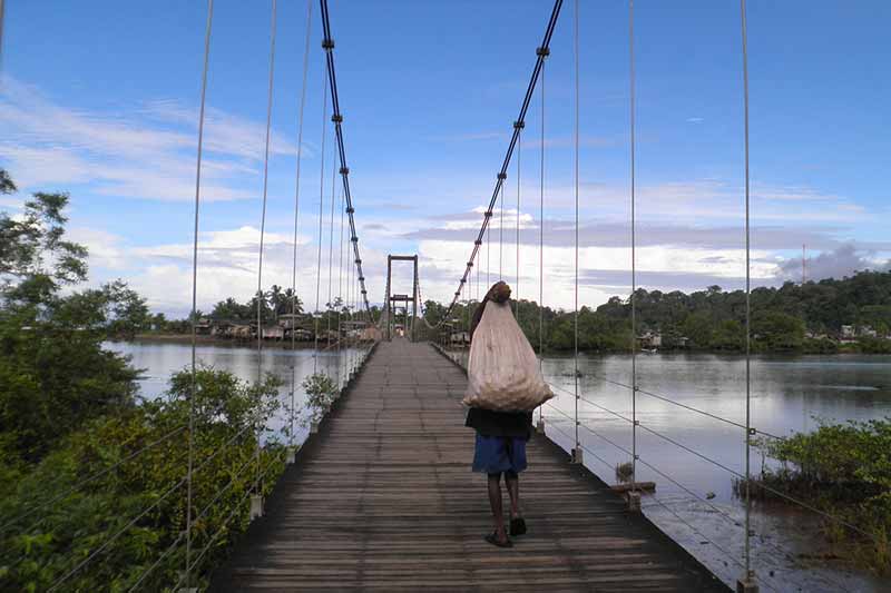 A young child holds a bag over their back and walks on a bridge	over a body of water.