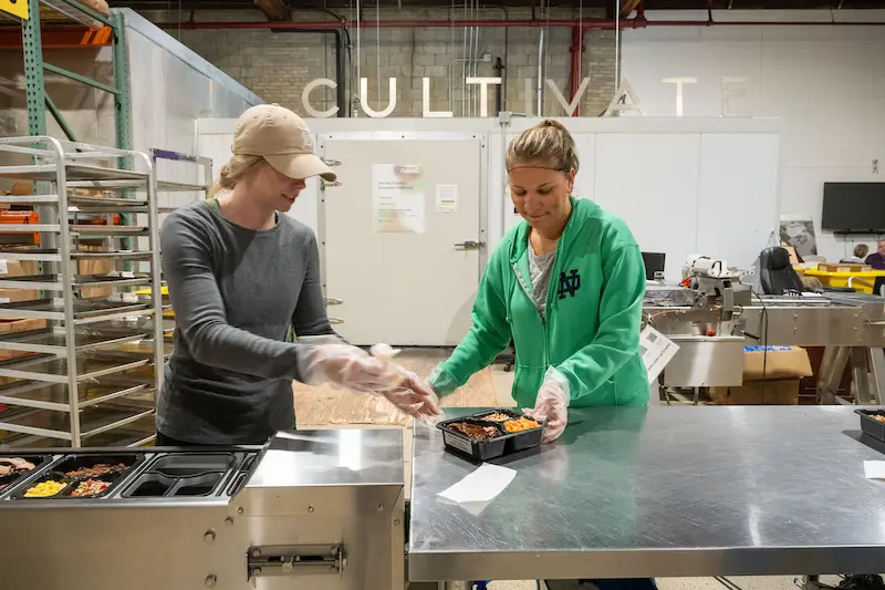 Two women, wearing gloves, package meals in a commercial kitchen with ”CULTIVATE“ on the wall and food trays in the background.