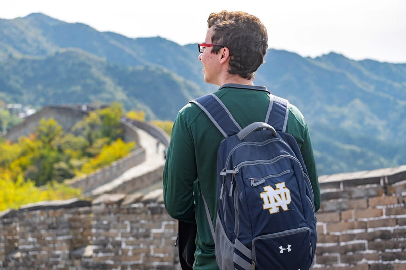 A male student with a Notre Dame backpack stands on a stone wall overlooking the Great Wall of China.A male student with a Notre Dame backpack stands on a stone wall overlooking the Great Wall of China.