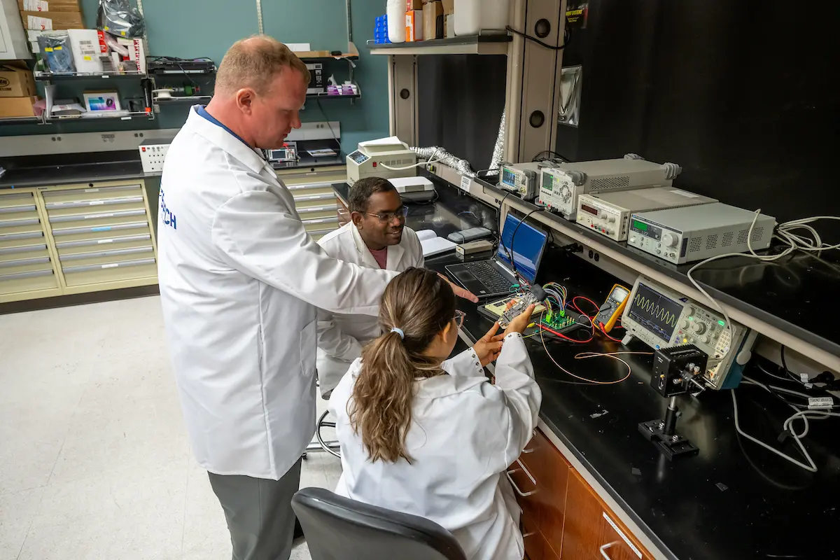 Three researchers in lab coats collaborate in a lab, working with electronic equipment and computers on a dark countertop.