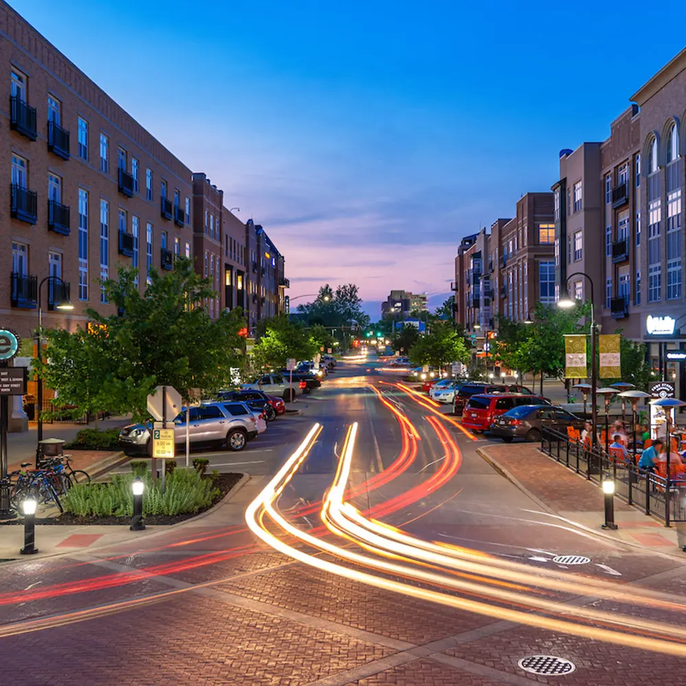 Long exposure shows Eddy Street Commons at dusk with light trails from cars on the street and illuminated buildings flanking both sides.